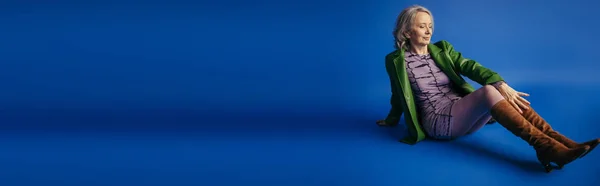 Full length of stylish grey haired woman in purple dress and green leather jacket sitting on blue background, banner — Stockfoto