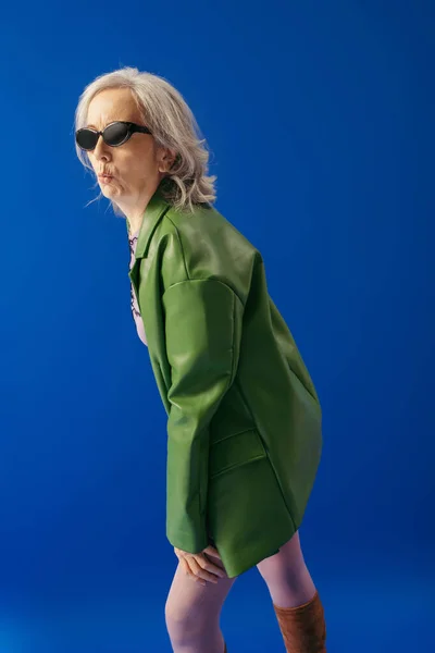 Grey haired woman in stylish sunglasses and green leather jacket pouting lips isolated on blue — Stock Photo