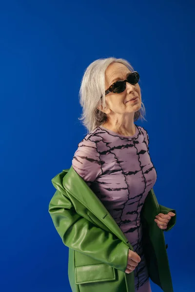 Senior woman in sunglasses and green leather jacket over purple dress smiling isolated on blue — Stock Photo
