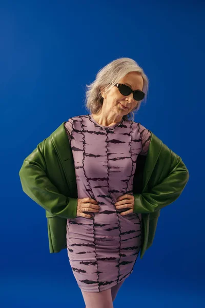 Smiling senior model in sunglasses and green leather jacket over purple dress posing with hands on waist isolated on blue — Stock Photo