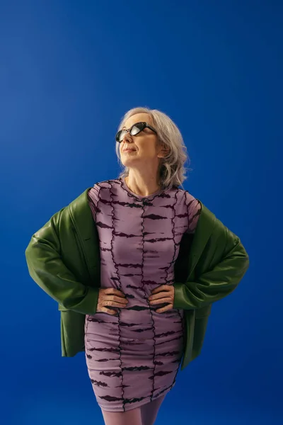 Senior model in sunglasses and fashionable outfit posing with hands on waist isolated on blue — Stock Photo