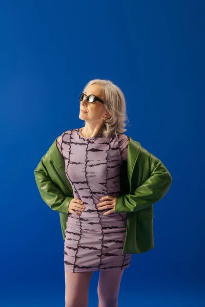 Stylish senior model in sunglasses and purple dress with green leather jacket standing with hands on waist isolated on blue — Stock Photo