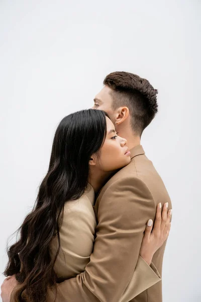 Вид збоку брюнетки азіатська жінка з довгим волоссям, що обіймає стильного чоловіка ізольовано на сірому — стокове фото