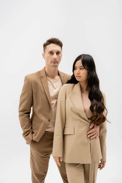 Uomo in abito beige in piedi con mano in tasca e abbracciando donna asiatica isolato su grigio — Foto stock