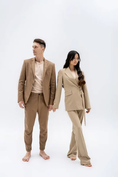 Full length of barefoot multiethnic couple in stylish pantsuits standing on grey background — Stockfoto