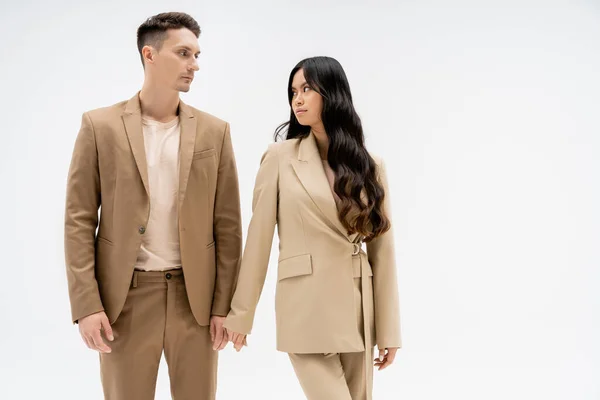 Fashionable multiethnic couple in beige suits looking at each other isolated on grey — Stock Photo
