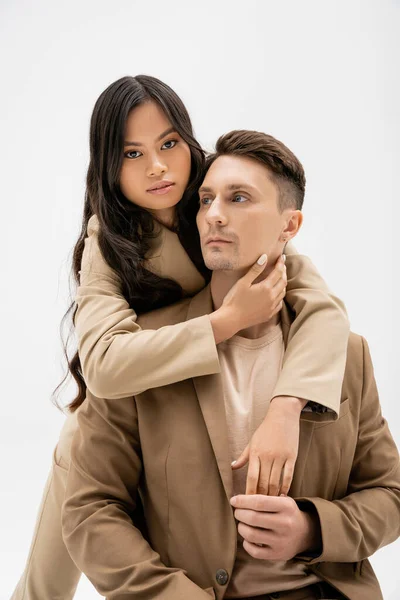 Брюнетка азиатка обнимает стильного мужчину в бежевом пиджаке и смотрит на камеру, изолированную на сером — стоковое фото