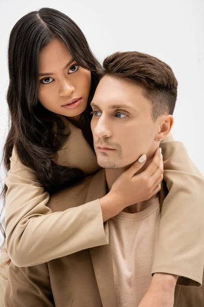 Брюнетка азіатська жінка дивиться на камеру і обіймає чоловіка в бежевому блістері ізольовано на сірому — стокове фото