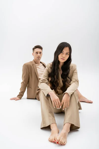 Barefoot asian woman with long brunette hair looking at camera near man in beige suit sitting on grey background — Stock Photo