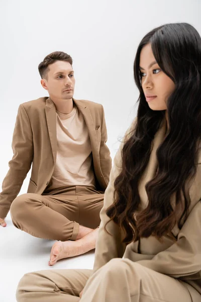 Asian woman with long brunette hair near barefoot man in beige suit sitting on grey background — Stock Photo