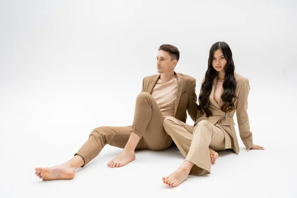 Full length of barefoot interracial couple in beige suits sitting on grey background — Stock Photo
