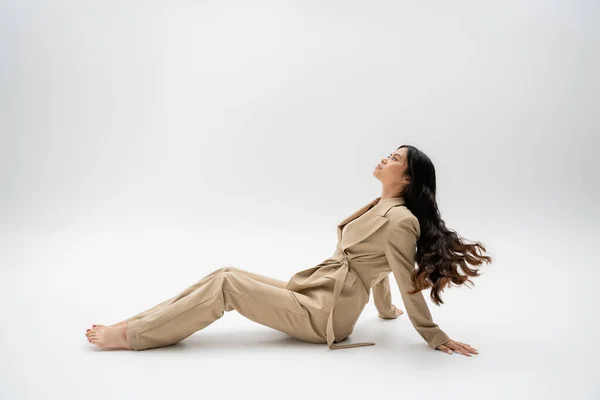 Vue latérale du modèle asiatique pieds nus avec des cheveux longs assis dans une combinaison à la mode sur fond gris — Photo de stock