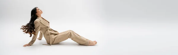 Side view of barefoot asian woman with long brunette hair sitting in beige suit on grey background, banner — Stock Photo