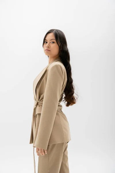 Jeune asiatique femme en beige veste regarder caméra tout en se tenant isolé sur gris — Photo de stock