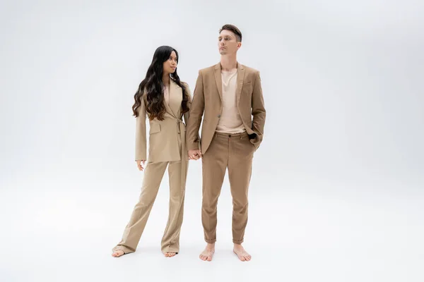Comprimento total de casal multiétnico descalço em ternos da moda de mãos dadas enquanto em pé sobre fundo cinza — Fotografia de Stock