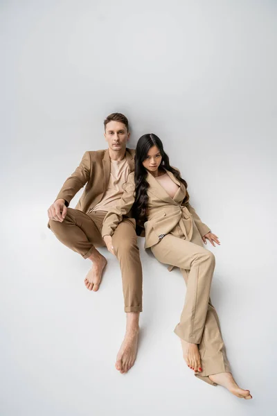 Full length of barefoot interracial couple in trendy outfit looking at camera while posing on grey background — Stock Photo