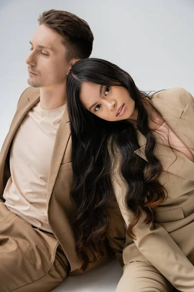 Morena mujer asiática con el pelo largo mirando a la cámara mientras está sentado cerca de hombre en traje beige sobre fondo gris - foto de stock
