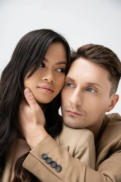 Trendy man in beige blazer embracing sensual asian woman isolated on grey — Stock Photo