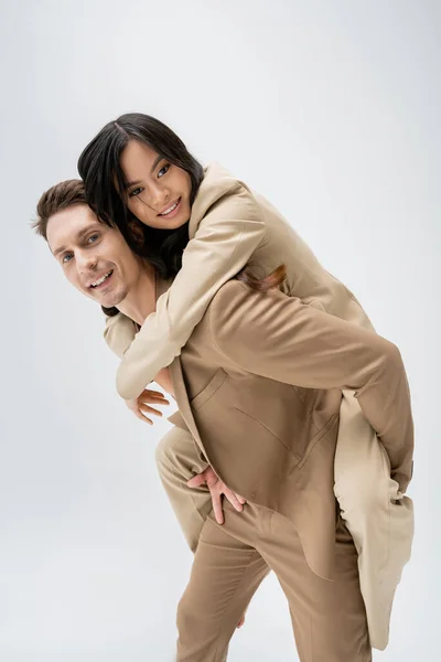 Happy and stylish man piggybacking young asian woman while smiling at camera isolated on grey — Stock Photo