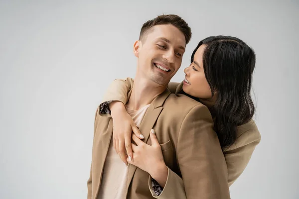 Brunette asian woman embracing happy man in beige stylish blazer isolated on grey — Stock Photo