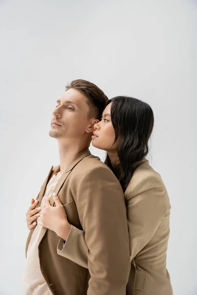 Sinnliche asiatische Frau umarmt Mann im trendigen Blazer isoliert auf grau — Stockfoto