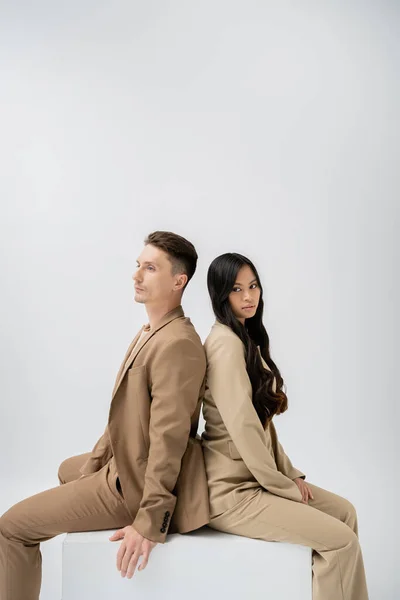 Brunette asian woman with long hair looking at camera while sitting back to back with trendy man isolated on grey — Stock Photo
