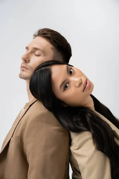Sensual asian woman looking at camera while leaning on man in beige blazer isolated on grey — Foto stock