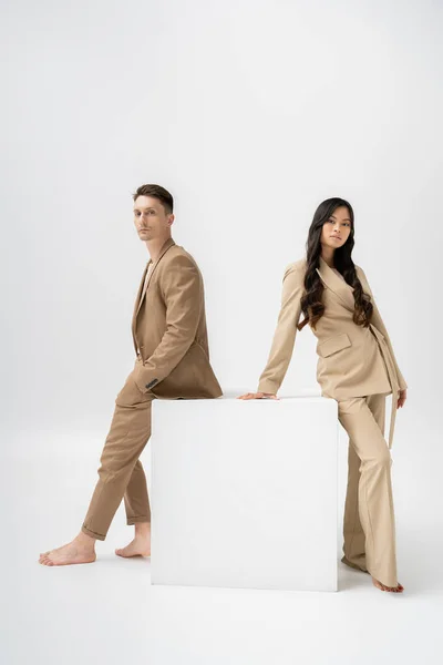 Full length of barefoot multiethnic couple in beige pantsuits looking at camera near white cube on grey background — Stock Photo