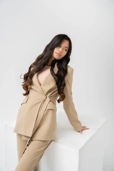 Charming asian woman in beige jacket and pants posing near white cube on grey background — Stock Photo