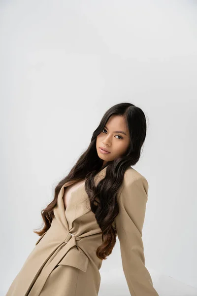 Pretty asian woman with long brunette hair posing in stylish blazer and looking at camera isolated on grey — Stockfoto