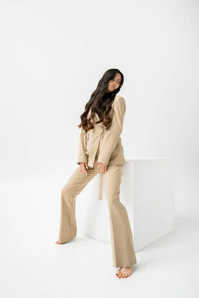 Full length of barefoot asian woman in beige suit sitting on cube on white background - foto de stock