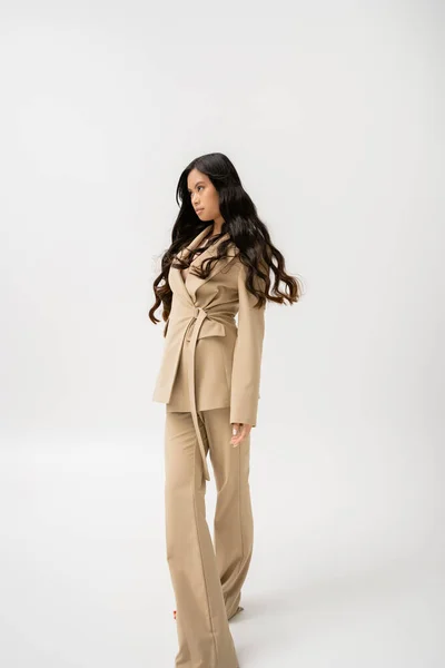 Long haired asian woman in beige pantsuit looking away while standing on grey background — Stockfoto