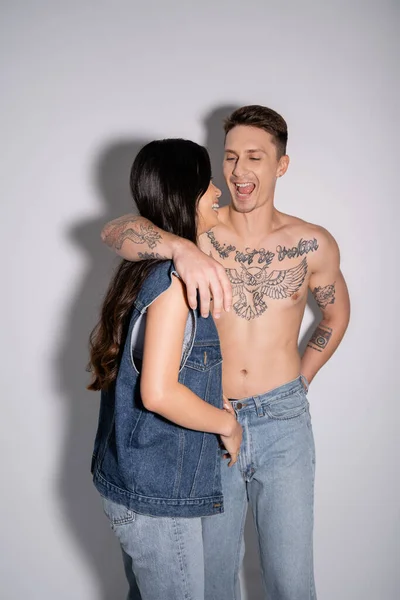 Excited shirtless man with tattooed body grimacing near asian woman in denim vest on grey background — Stock Photo