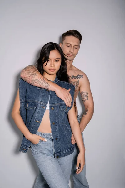 Hemdlos tätowierter Mann umarmt asiatische Frau posiert mit der Hand in der Hosentasche der Jeans auf grauem Hintergrund — Stockfoto