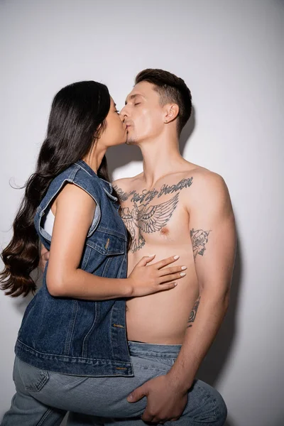 Brunette asian woman in denim vest kissing with shirtless tattooed man on grey background — Stockfoto