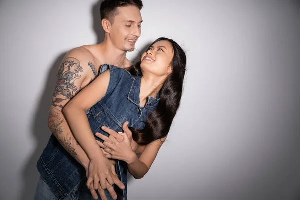 Torse nu tatoué homme étreignant heureux asiatique femme en denim gilet sur fond gris — Photo de stock