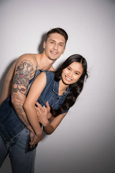 Tattooed and shirtless man embracing happy asian woman in denim vest on grey background — Stock Photo