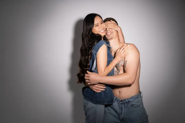 Cheerful asian woman in denim clothes covering eyes of shirtless tattooed man on grey background - foto de stock