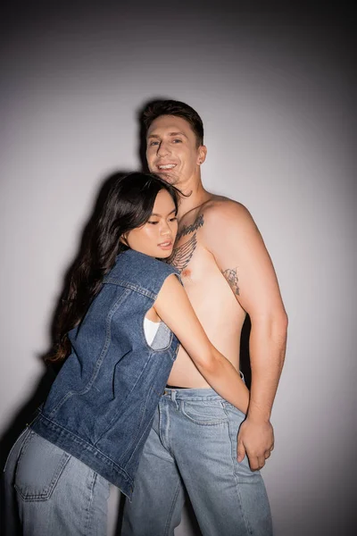Long haired asian woman in denim vest embracing shirtless tattooed man smiling at camera on grey background — Photo de stock