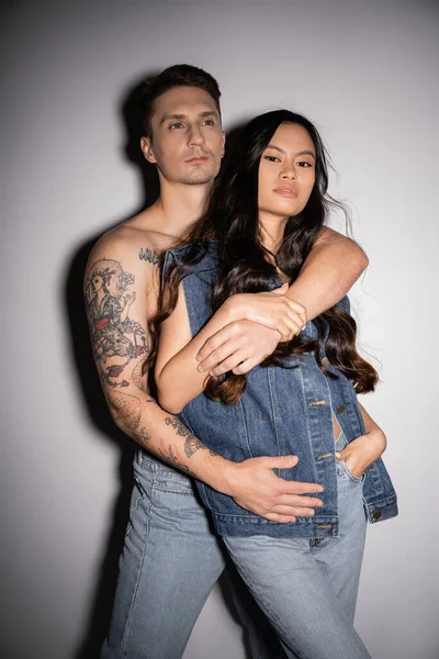 Shirtless tattooed man hugging brunette asian woman in denim clothes looking at camera on grey background — Stock Photo
