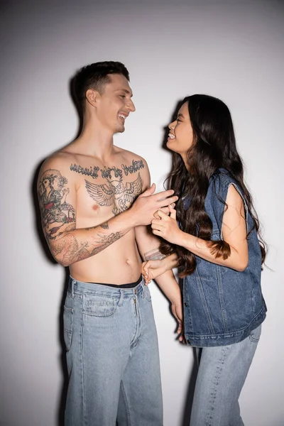 Cheerful asian woman and shirtless tattooed man smiling at each other on grey background - foto de stock