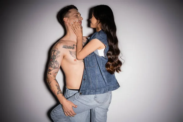 Smiling asian woman in denim clothes touching face of tattooed muscular man on grey background — Stock Photo