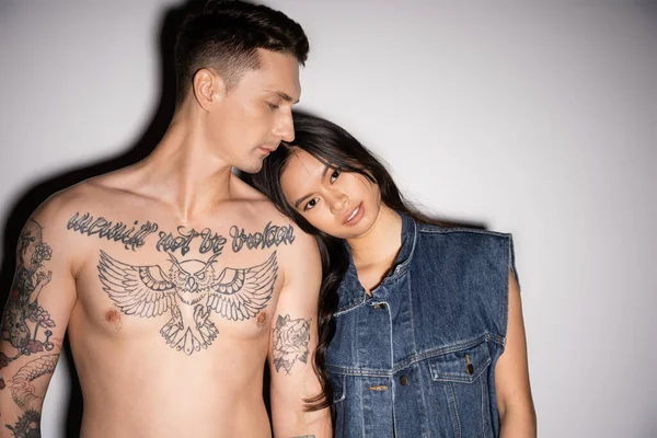 Asian woman in denim vest looking at camera while leaning on shirtless tattooed man on grey background - foto de stock