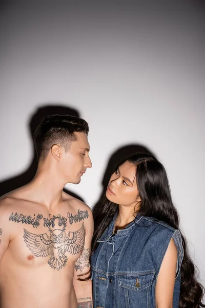 Shirtless man with tattooed body and brunette asian woman with long hair looking at each other on grey background — Stock Photo