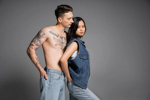 Brunette asian woman and tattooed shirtless man standing with hands in back pockets of jeans on grey background — Stock Photo