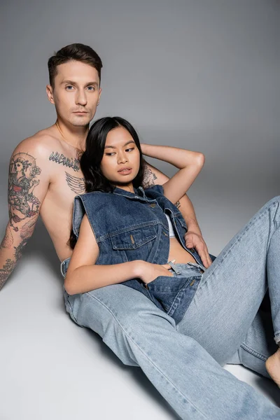 Tattooed shirtless man looking at camera while sitting near asian woman in denim clothes on grey background — Fotografia de Stock