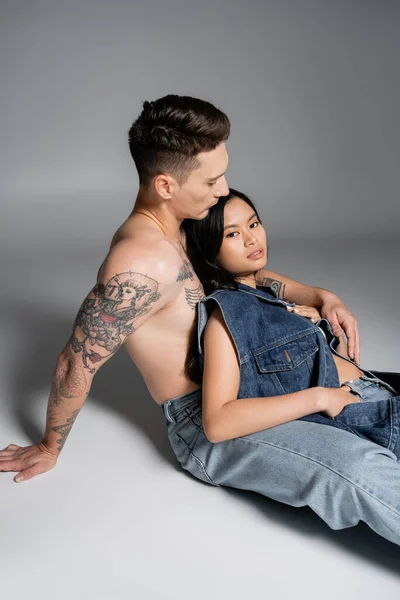Pretty asian woman in denim vest looking at camera while sitting near shirtless tattooed man on grey background - foto de stock
