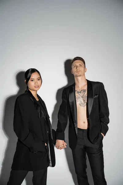 Stylish interracial couple in black elegant blazers holding hands and looking at camera on grey background — Stockfoto