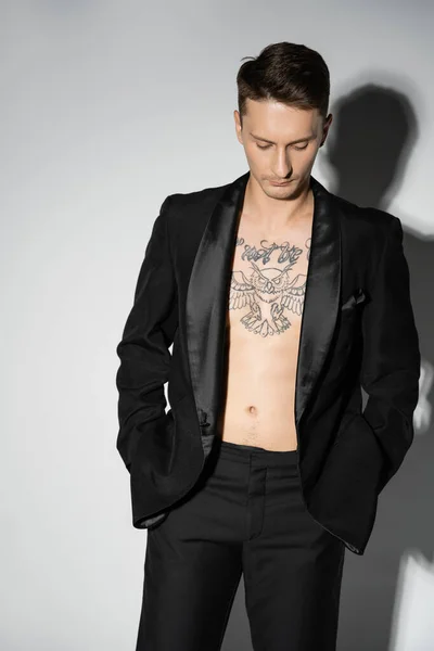 Модный мужчина с татуировками в черном пиджаке над телом без рубашки, держась за руки в карманах на сером фоне с тенью — стоковое фото