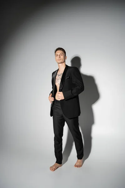 Full length of barefoot man in black elegant suit standing on grey background with shadow — Stock Photo